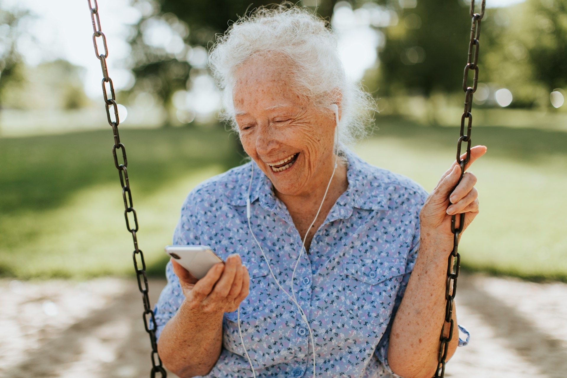 Elderly person smiling, using a smartphone while sitting on a swing set outdoors.
