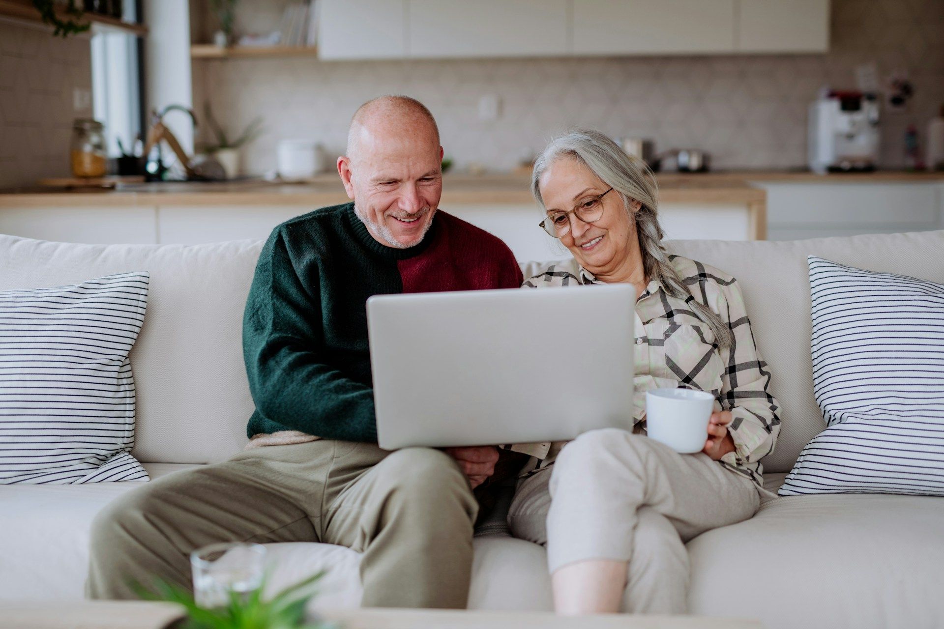 Couple on sofa looking at laptop, smiling. One holds a mug. Living room setting.