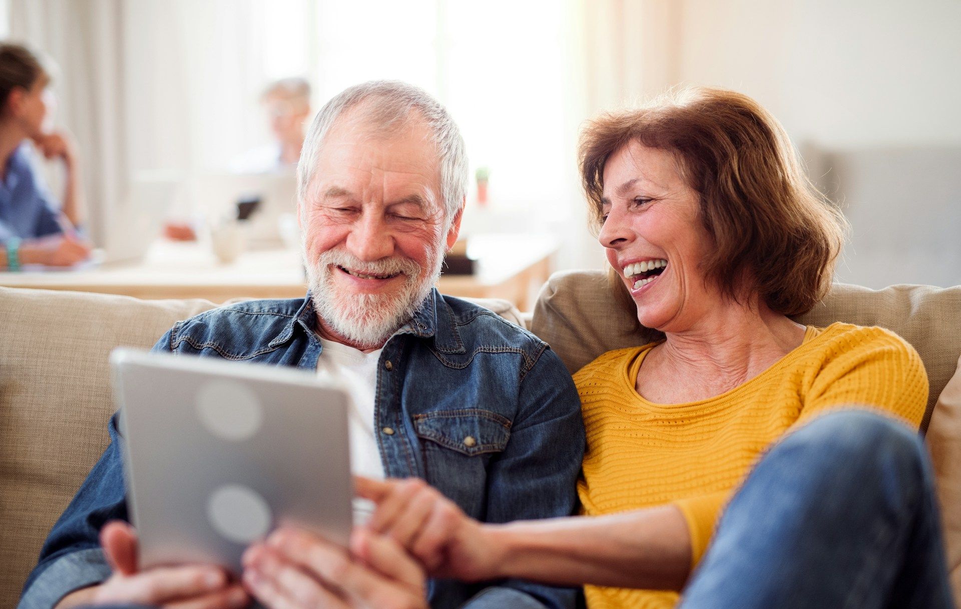 Elderly couple smiling at a tablet, seated on a couch indoors.