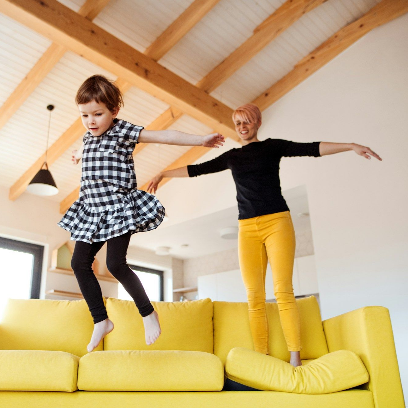 Woman and child jump on a yellow couch in a brightly lit living room; both have arms outstretched.