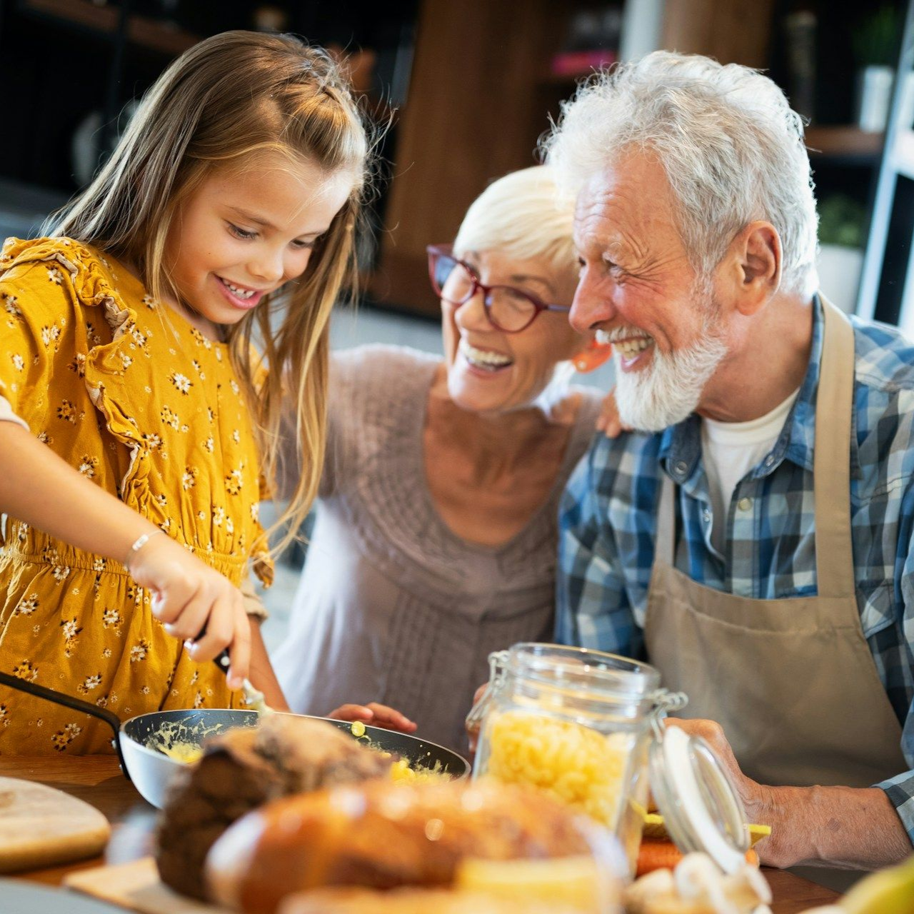 Girl cooking with grandparents; all smiling in a kitchen setting.