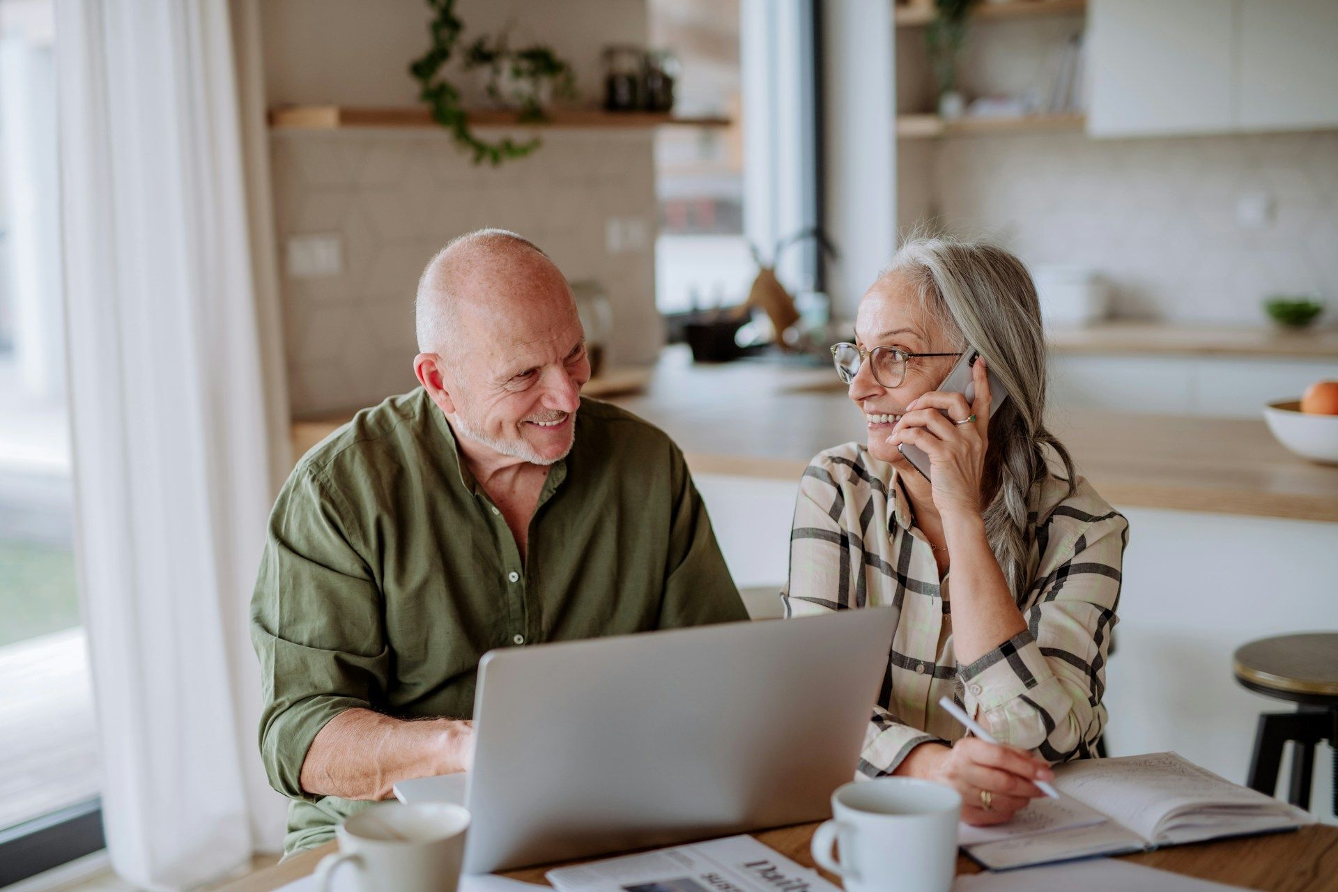 Older couple at table with laptop; woman on phone, man smiles. Bright kitchen setting.