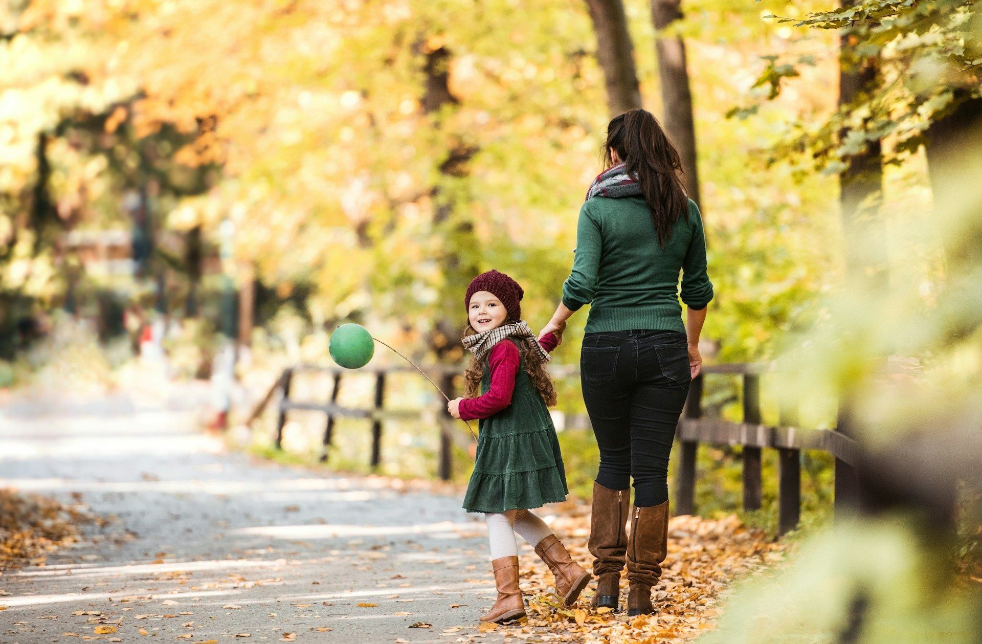 Woman and child walking on a path in autumn, holding hands. Child has a green balloon.