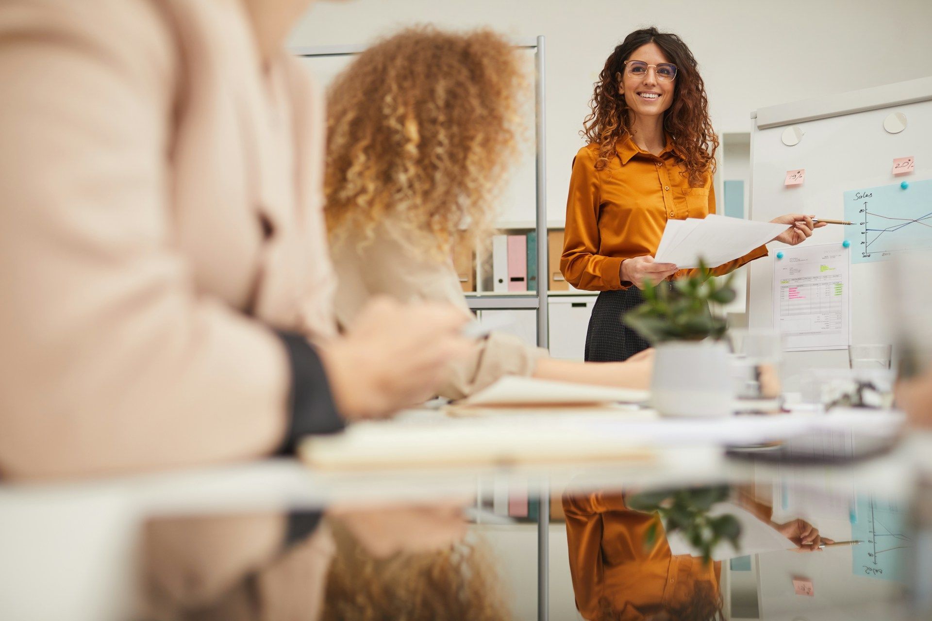 Woman in orange shirt presents at whiteboard, pointing to a diagram; colleagues at table.