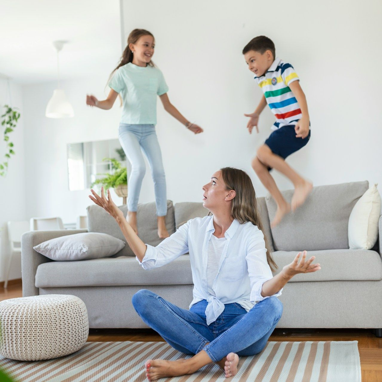 Woman seated on floor watches two children jumping on a couch indoors.
