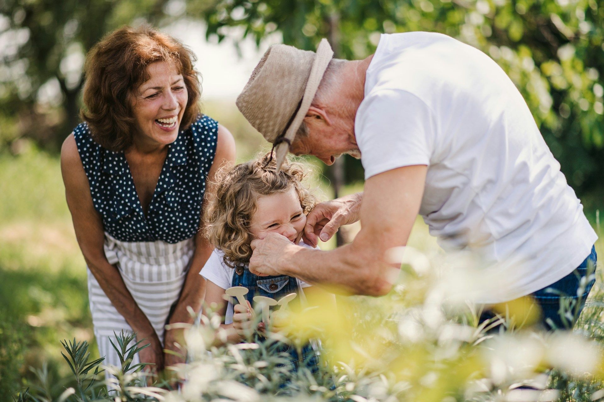 Grandparents playing with a child in a garden; woman laughs, man adjusts child’s hair.