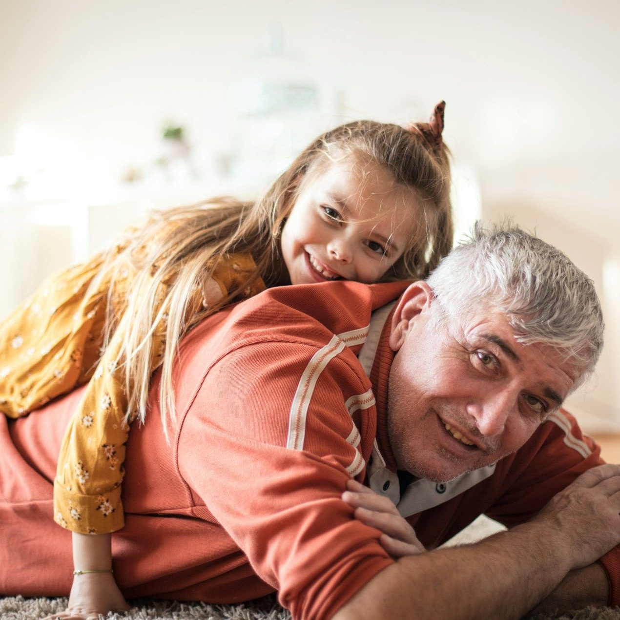 Girl with long hair on the back of a man in orange shirt, both smiling on the floor.