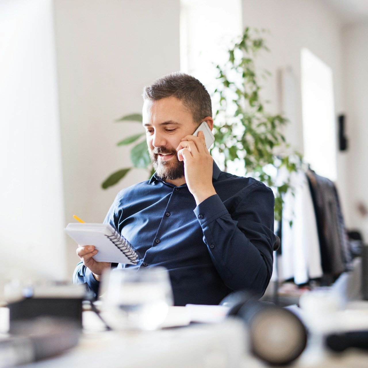 Man in blue shirt, on the phone, smiling, taking notes in a notebook at an office desk.