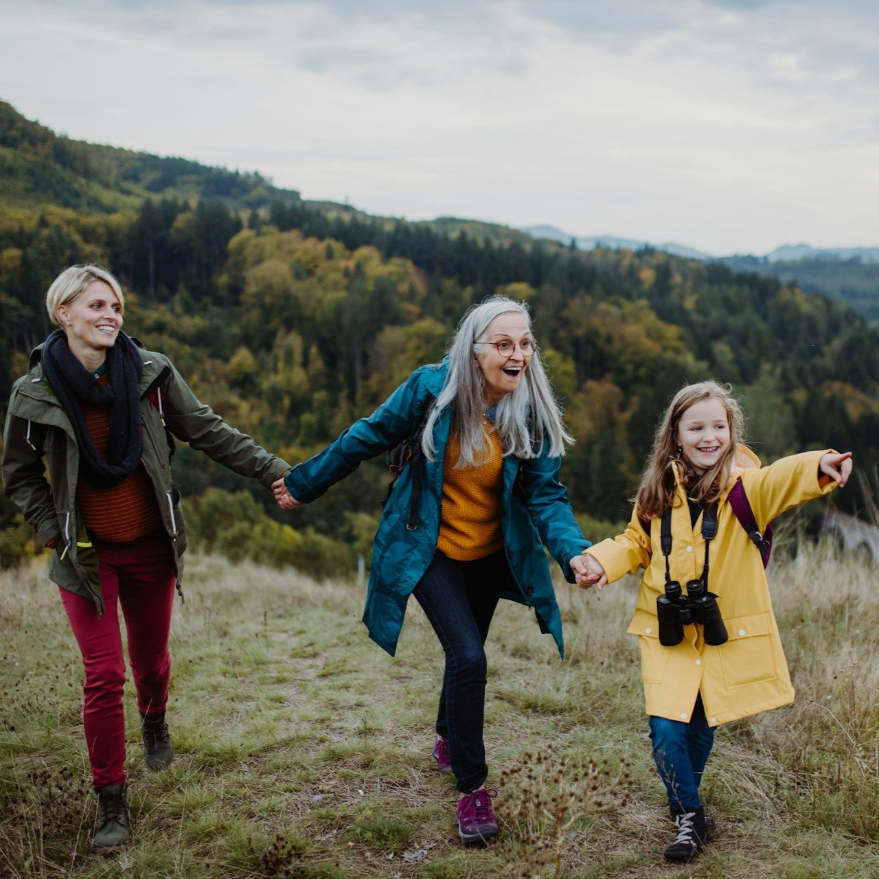 Three people hiking: a woman, older woman, and child holding hands, smiling, in front of a green mountain.