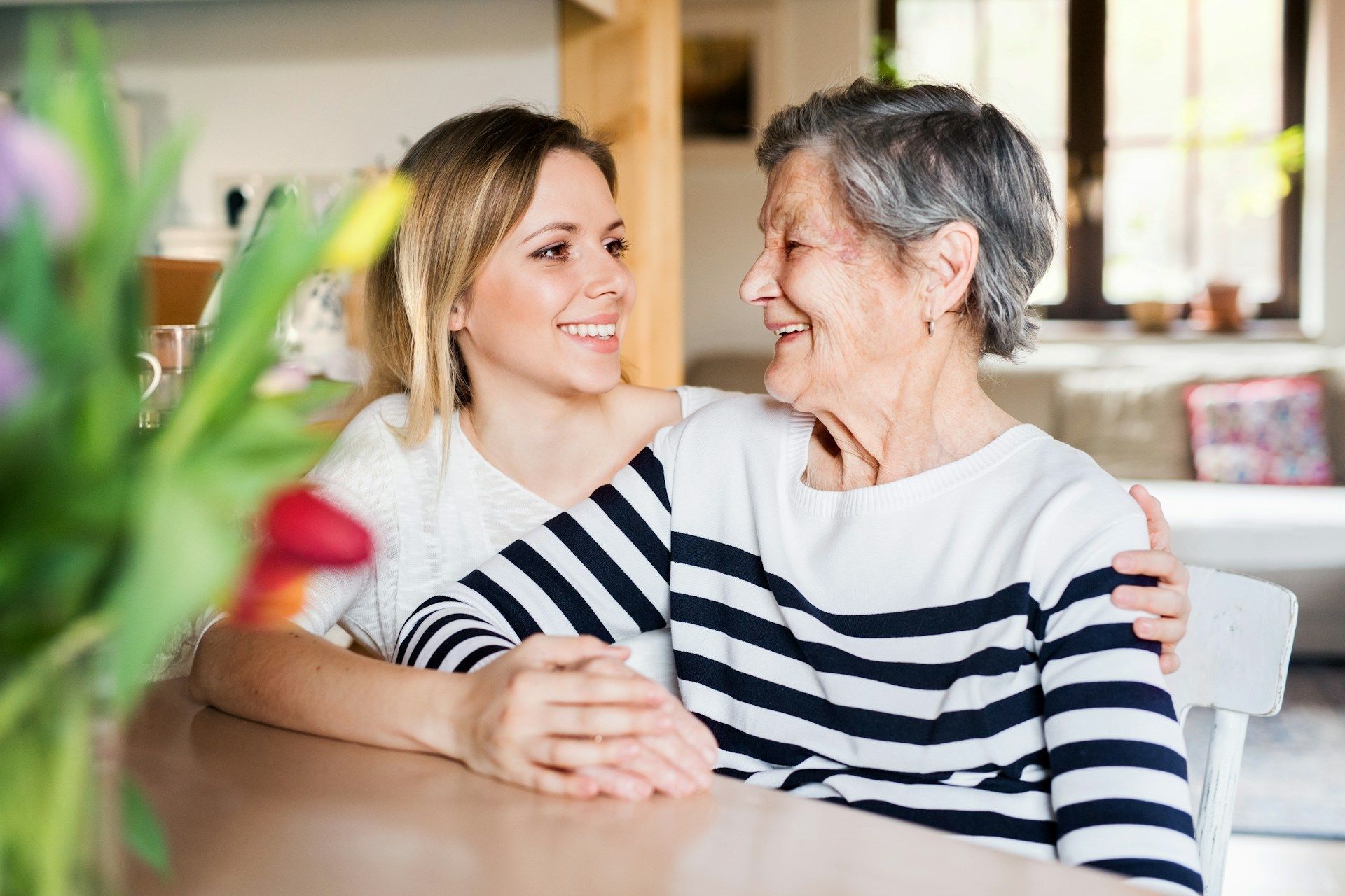 Young woman with arm around older woman, both smiling at each other at a table near flowers.