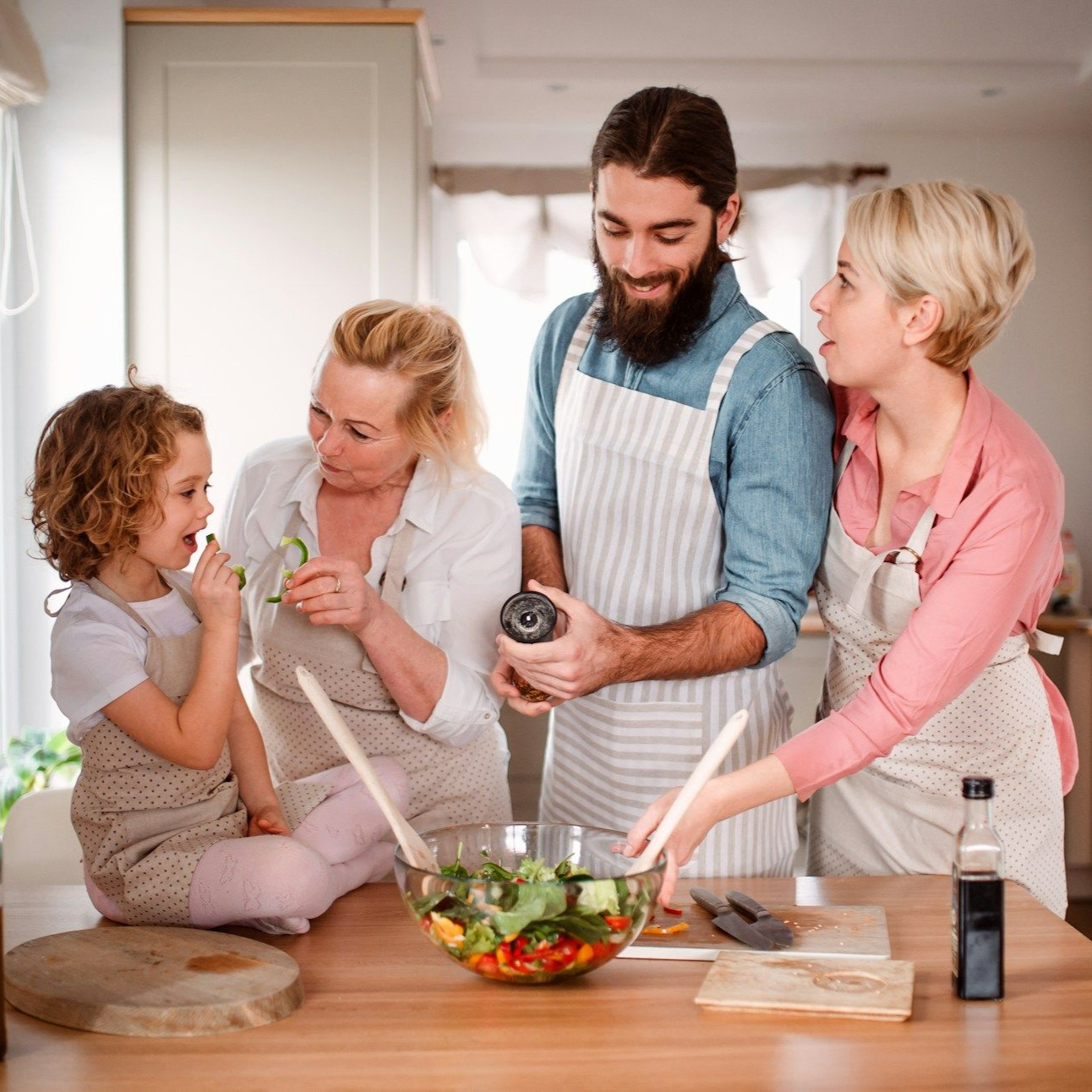 Family in kitchen cooking salad; child, adult, and older adult sharing ingredients, smiling.