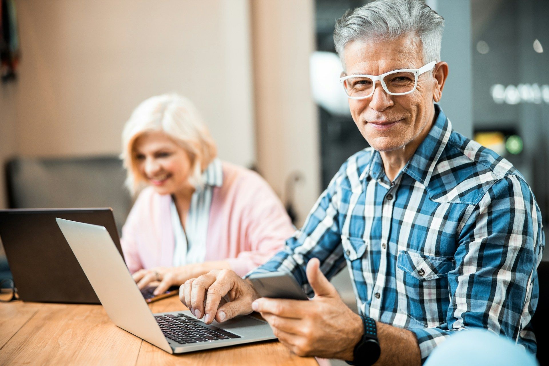 Man with glasses using laptop and phone, smiling. Woman in background using laptop. Cafe setting.