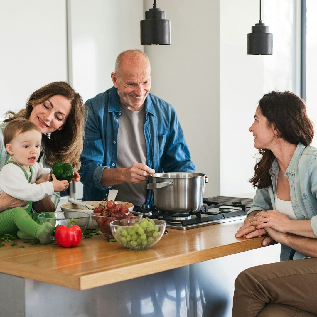 Family cooking together in a kitchen. Baby held by woman, man stirring pot, woman smiling.