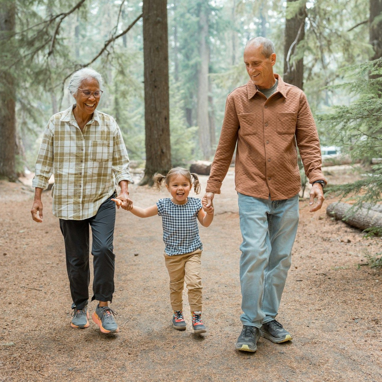 A child holds hands with two adults on a forest path. All are smiling and walking forward.