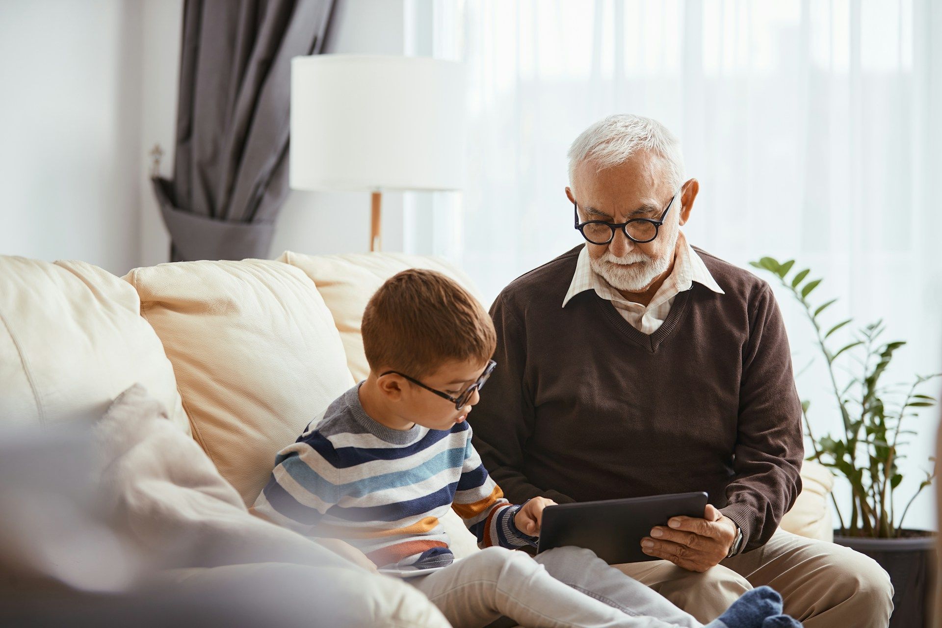 Grandfather and grandson sitting on a couch, looking at a tablet together in a living room.