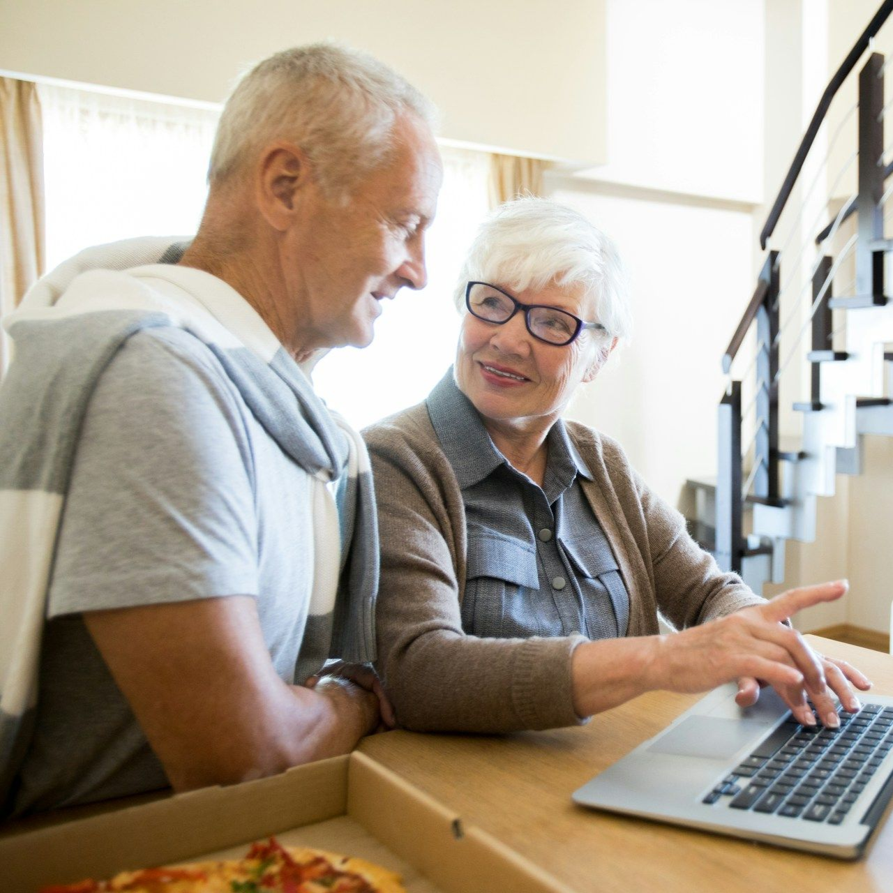 Elderly couple using a laptop, smiling. Pizza box on the table.