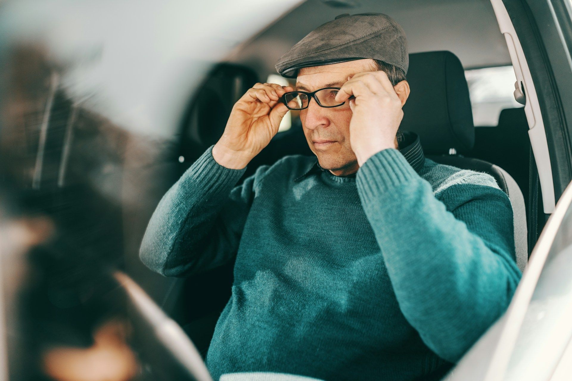Man inside a car adjusting glasses, wearing a cap and blue sweater.