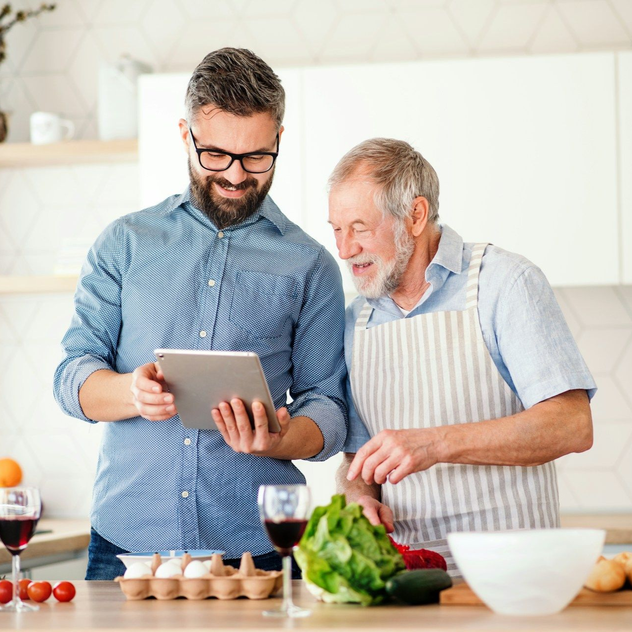 A man and older adult looking at a tablet while cooking in a kitchen.