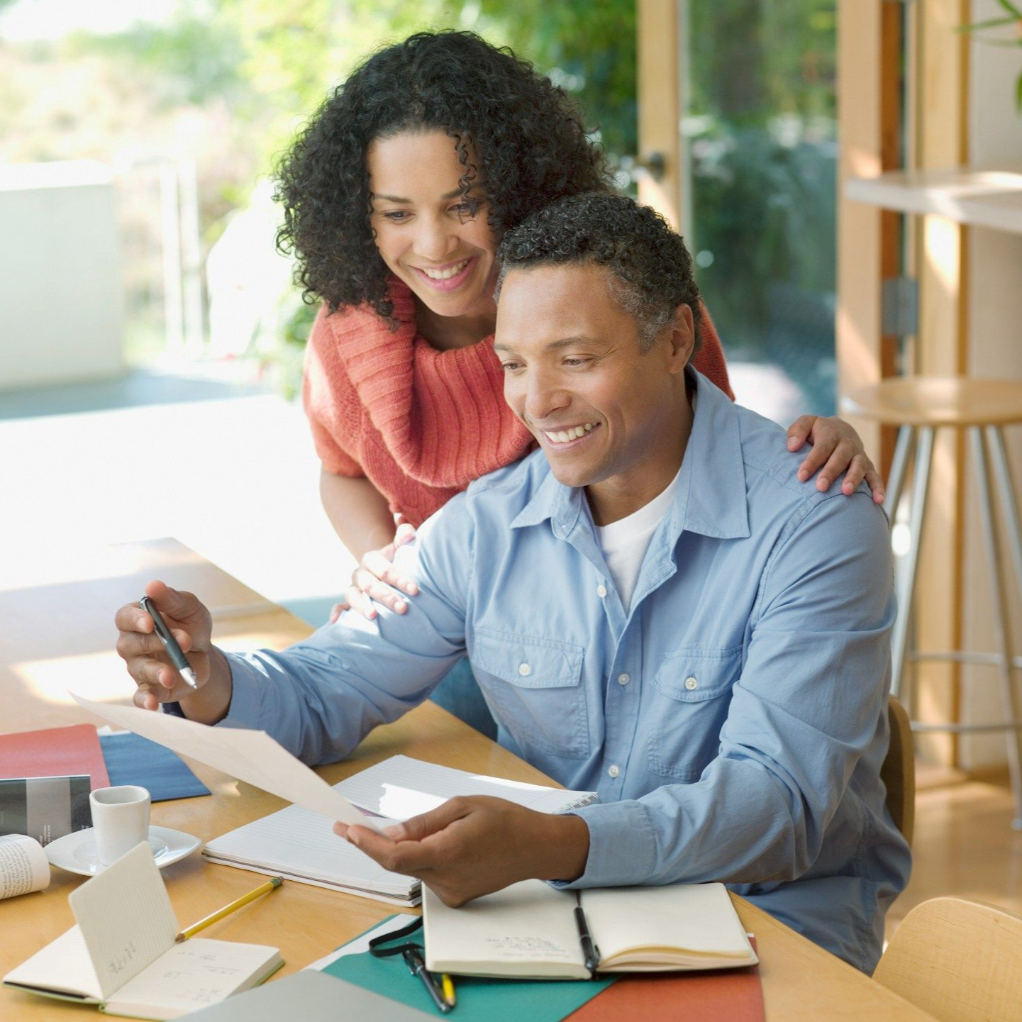 Couple smiling, looking at papers at a table. Woman has arm on man's shoulder. Natural light in room.