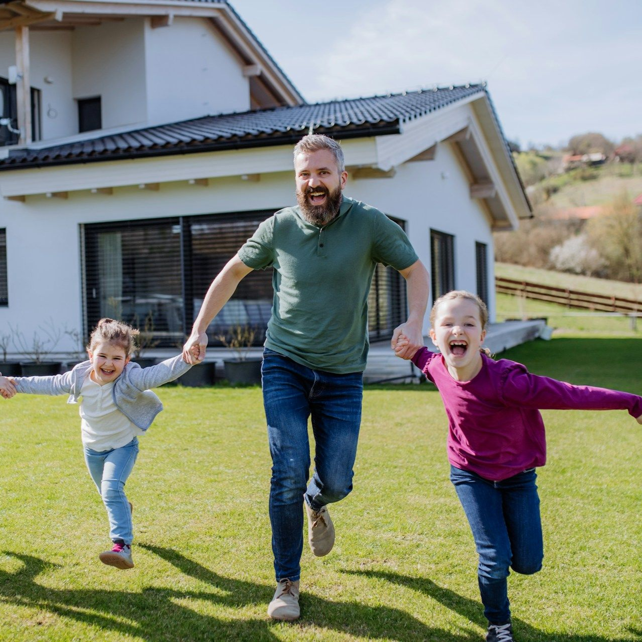 Man and two children running on a lawn in front of a house, all smiling.