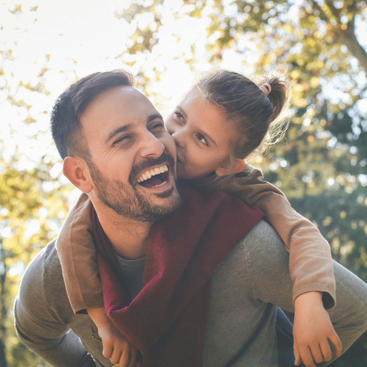 Man smiling, carrying a child on his back, who is kissing his cheek; outdoors.