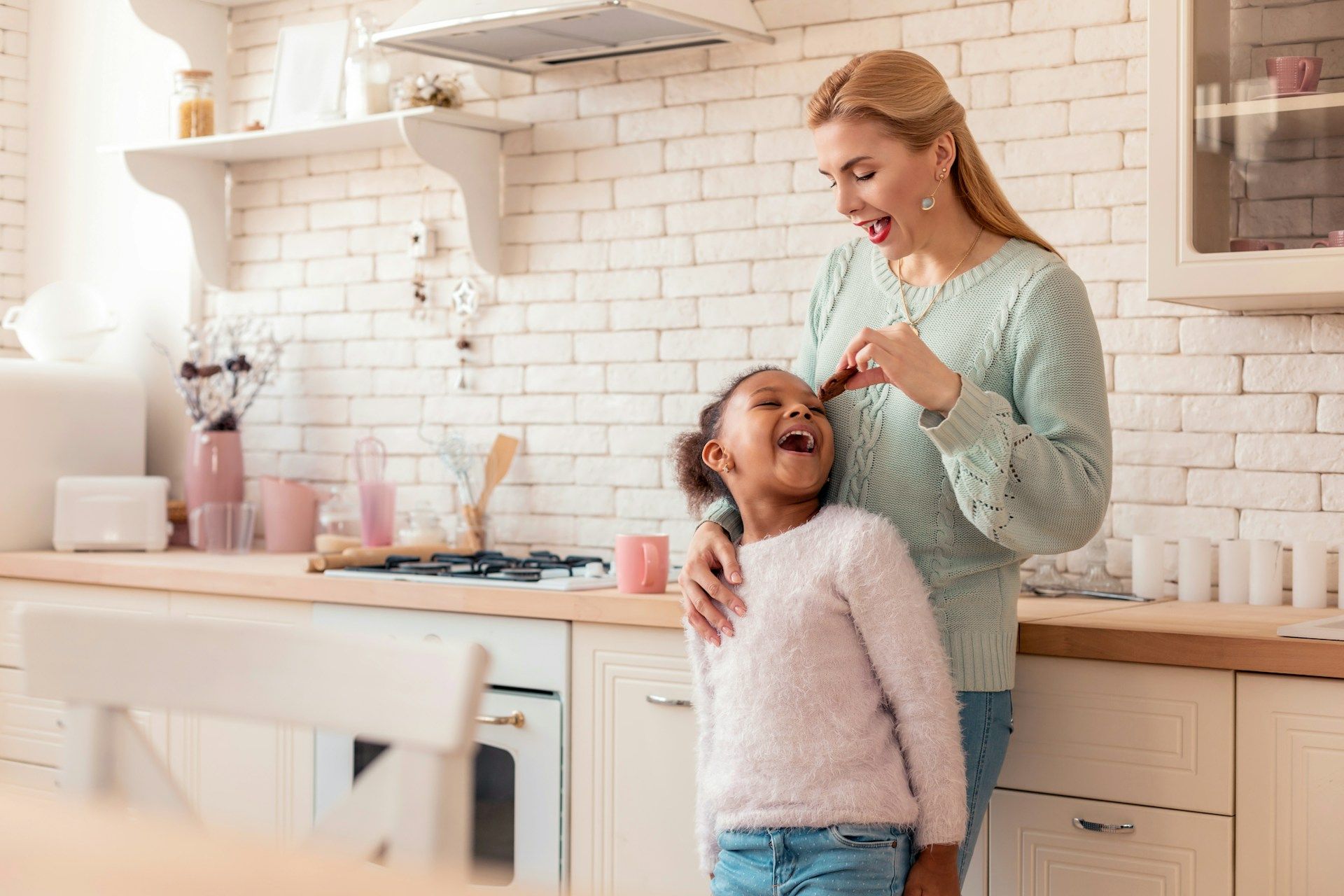 Woman combing a child's hair in a bright kitchen; both smiling and laughing.