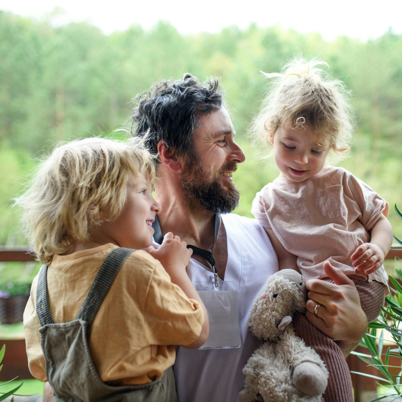 Man holding two children on a balcony, smiling. The children look at him, one holds a stuffed animal.