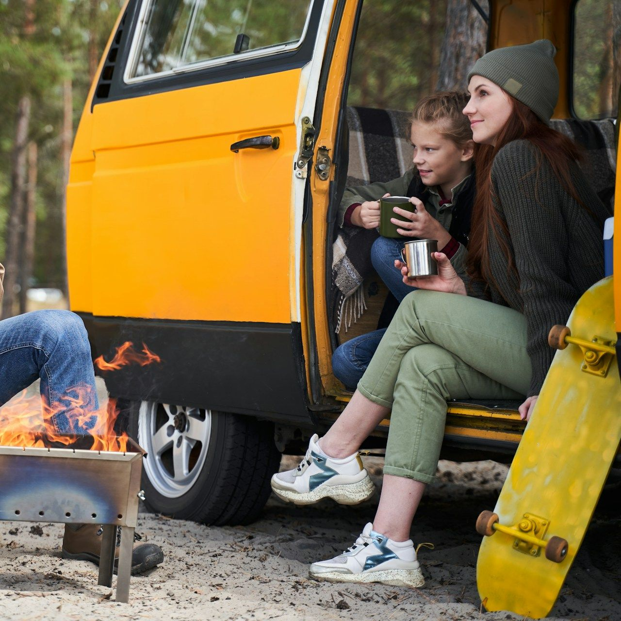 Woman and child sit in a yellow van, near a campfire and a skateboard.