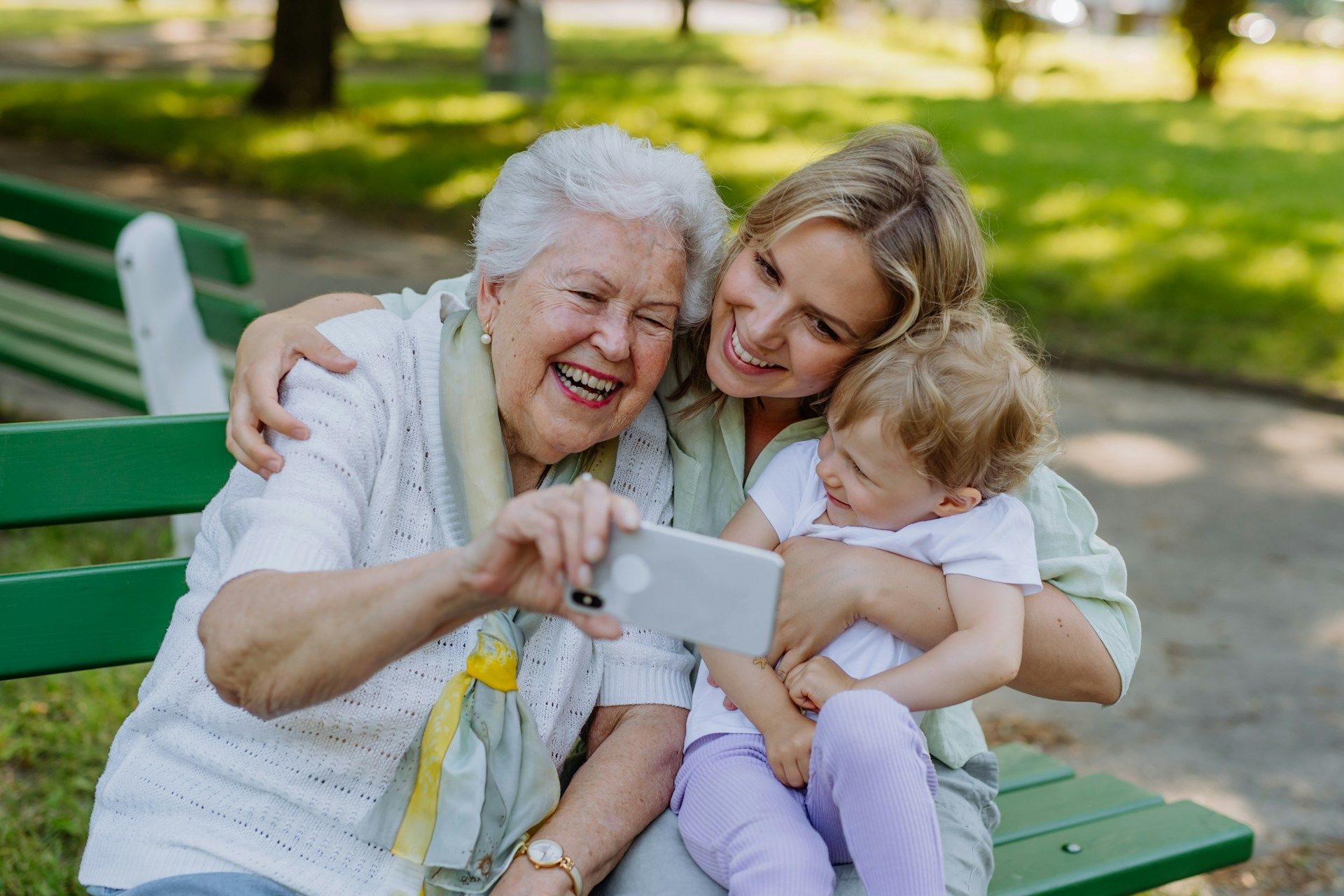 Grandmother, mother, and child take a selfie on a park bench, all smiling.