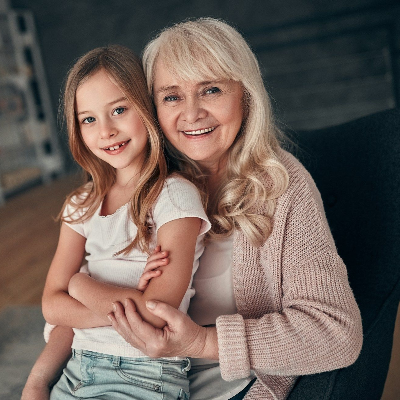 A smiling girl with a missing tooth hugs an elderly woman seated in a chair. Both look at the camera.
