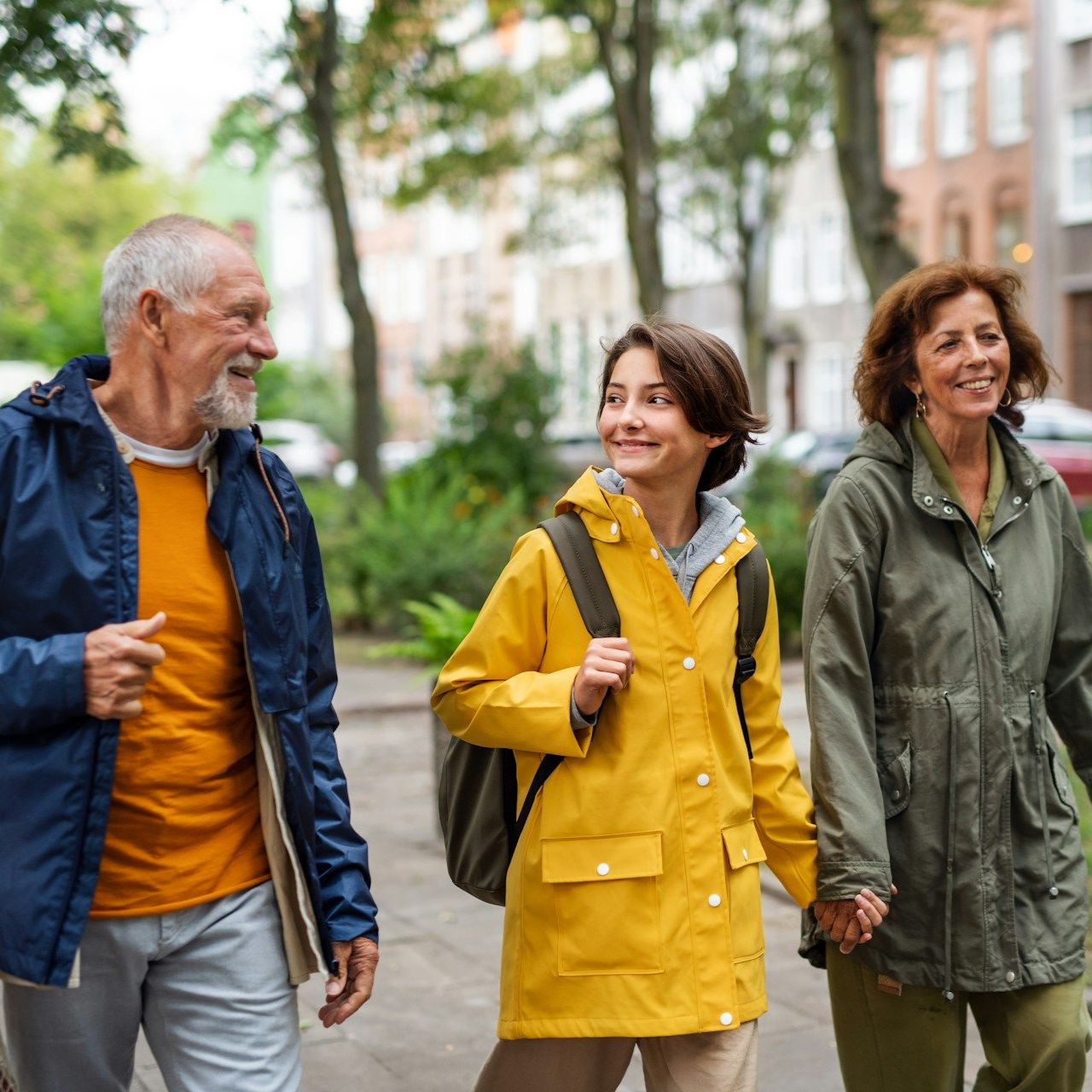 A child in a yellow raincoat walks between two adults on a sidewalk, smiling. Buildings and trees in background.
