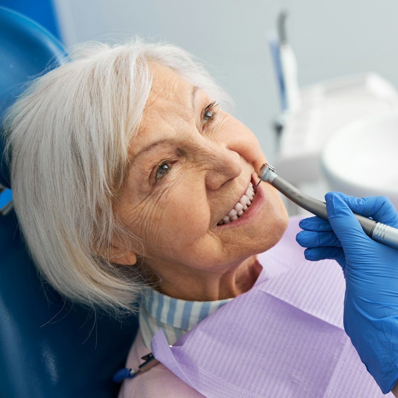 Woman in dental chair, smiling, receiving dental work. Blue gloved hand holds tool near teeth.