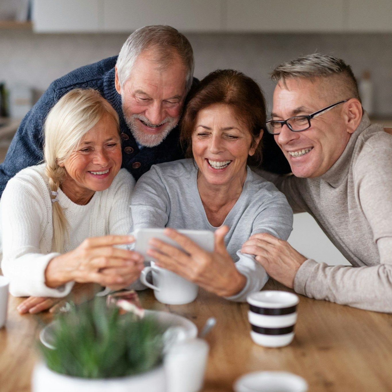 Four people looking at a tablet together, smiling. Inside a kitchen, near a table.