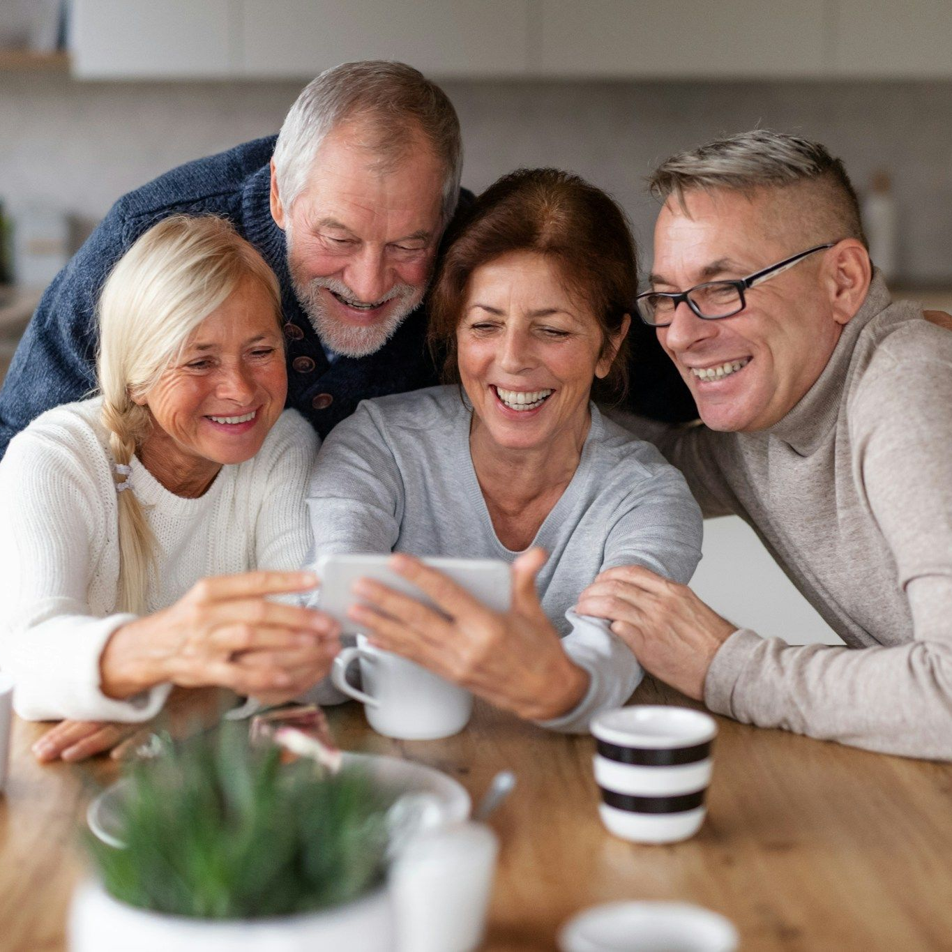 Four people smiling, gathered around a tablet at a table, likely looking at a screen in a kitchen.