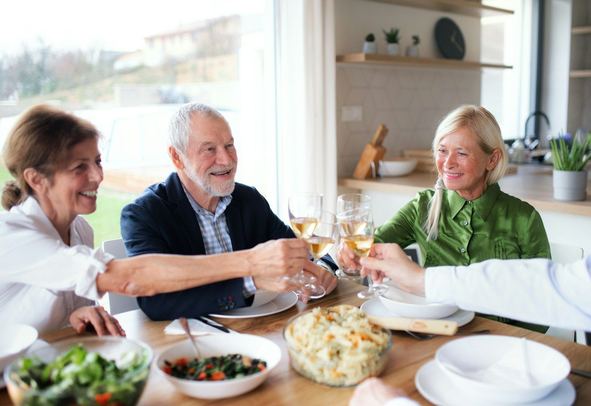 Four people toasting drinks at a dining table. Sunlight illuminates the scene, with a meal and greenery present.