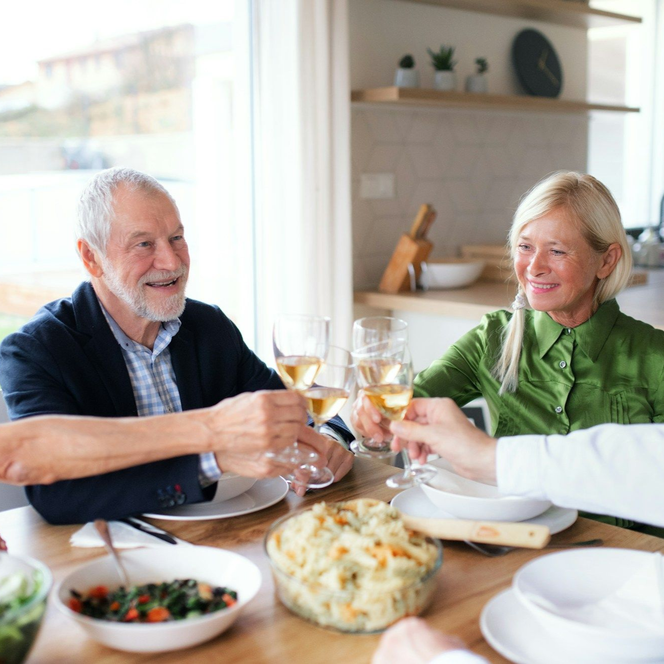 Two people, one with a beard, toasting with wine at a dining table with others.
