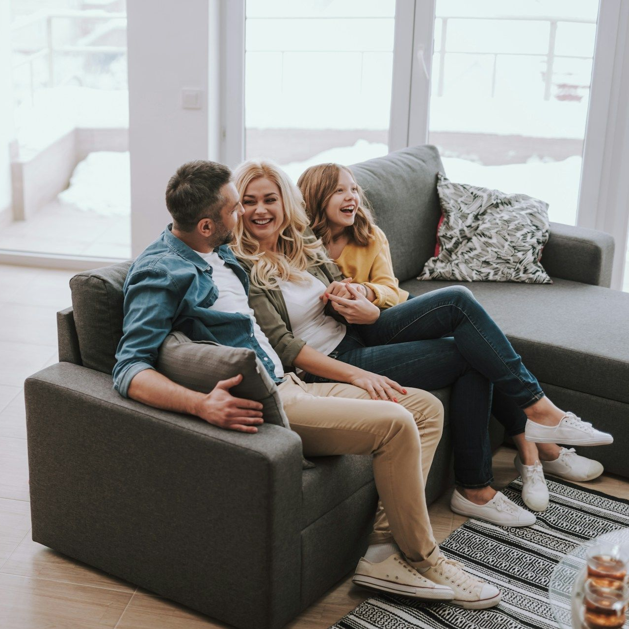 Family of three laughing on a gray couch in a living room with large windows.