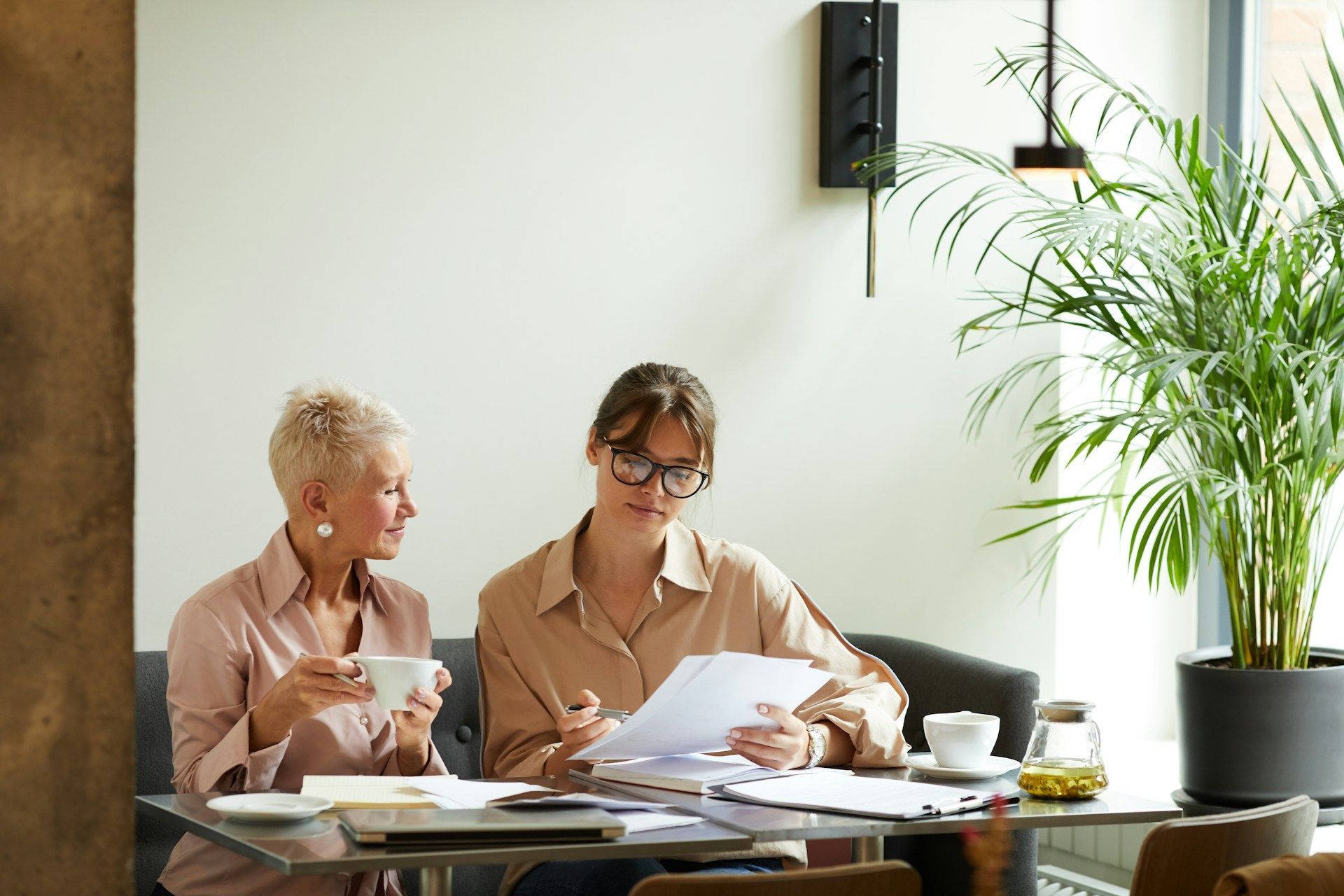 Two women reviewing documents at a table. One holds a cup, the other wears glasses. A potted plant is nearby.