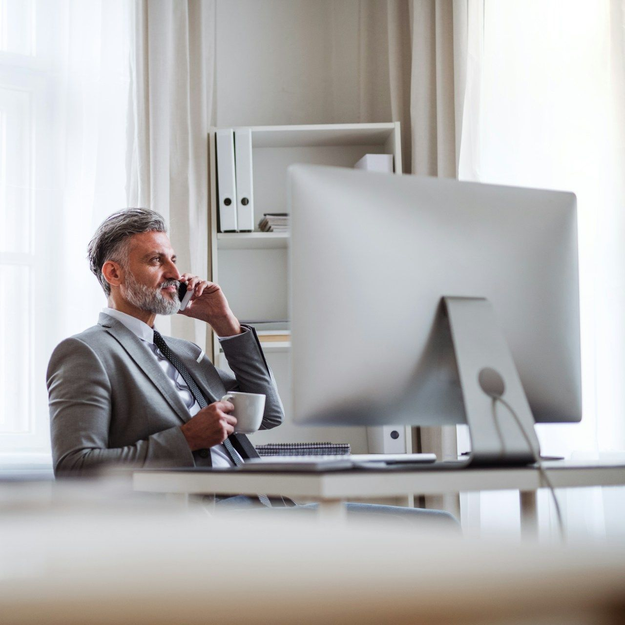 Man in suit, on phone, holding mug, at desk with computer in office setting.