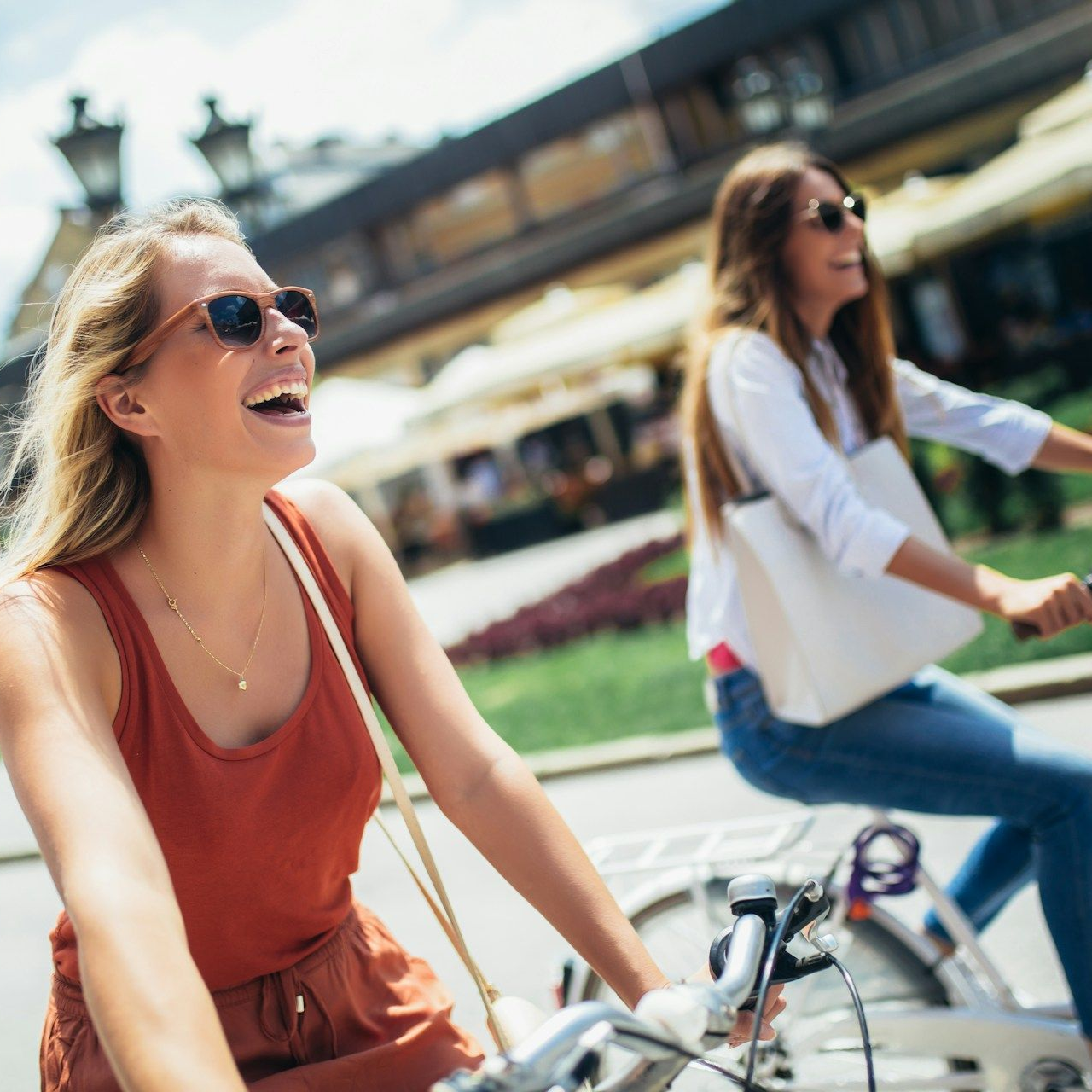 Two women riding bikes, smiling in sunny city. Woman in red top laughs, other wears white shirt.