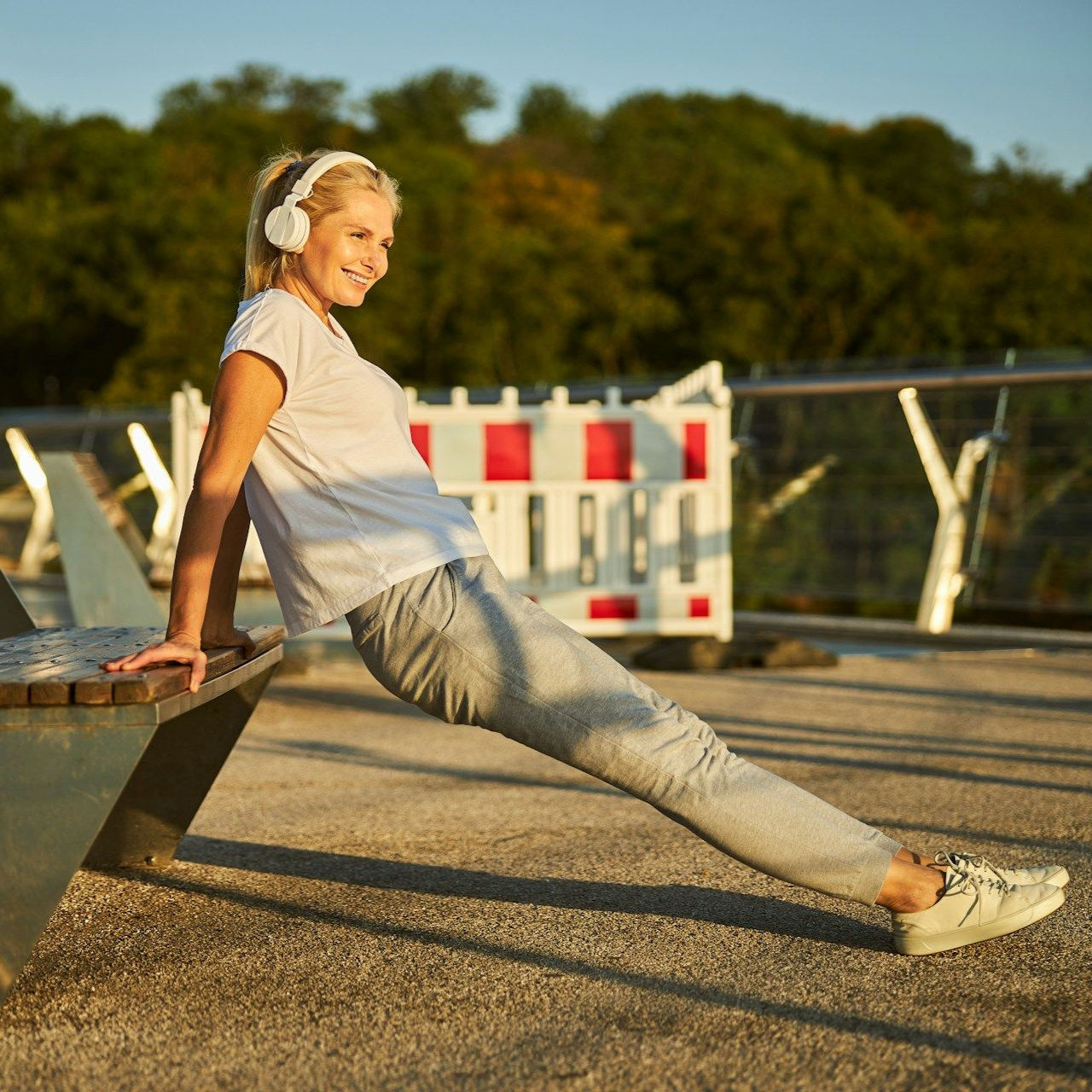 Woman in white shirt and grey sweatpants doing tricep dips on a bench outdoors.