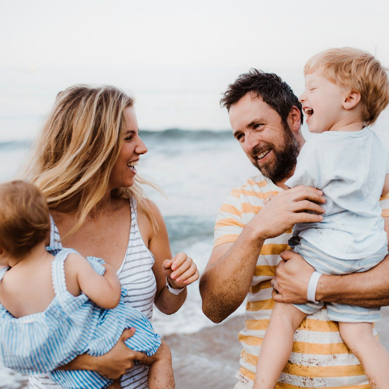 Family laughing on a beach; woman holding a toddler, man holding a boy. Ocean in background.