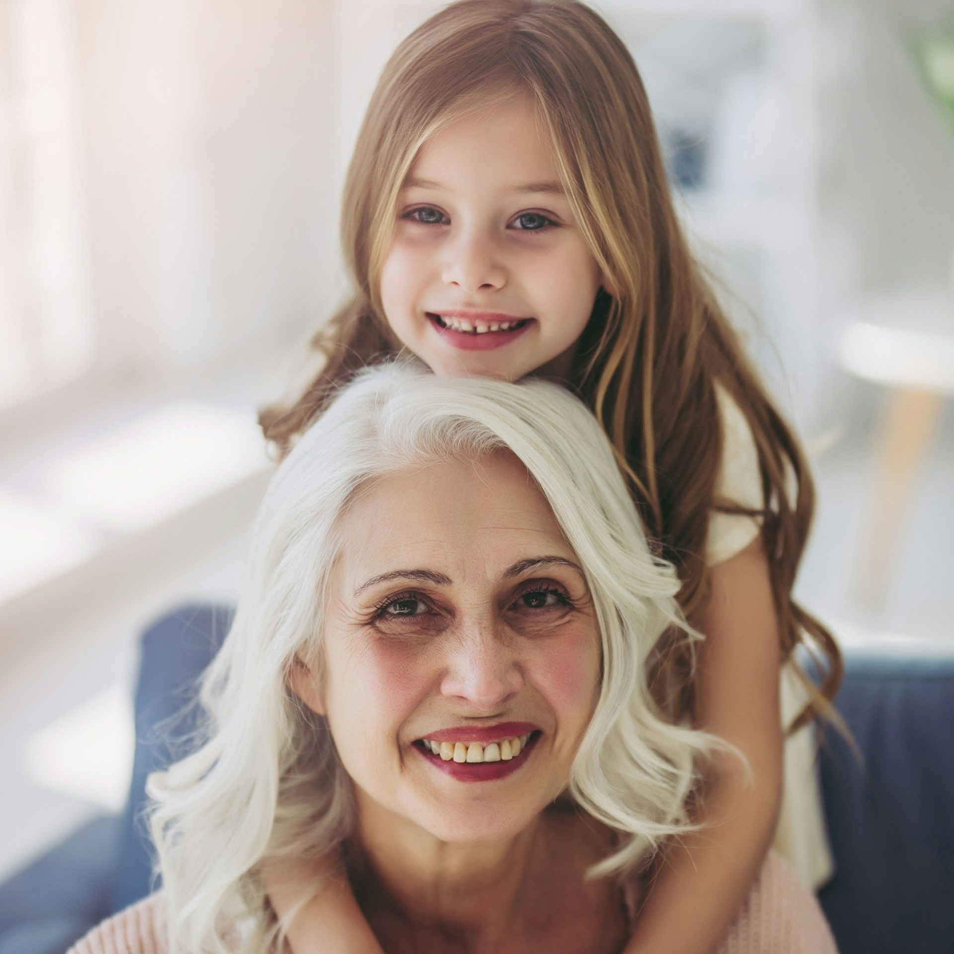 Young girl with long hair hugs a woman with white hair from behind, both smiling.