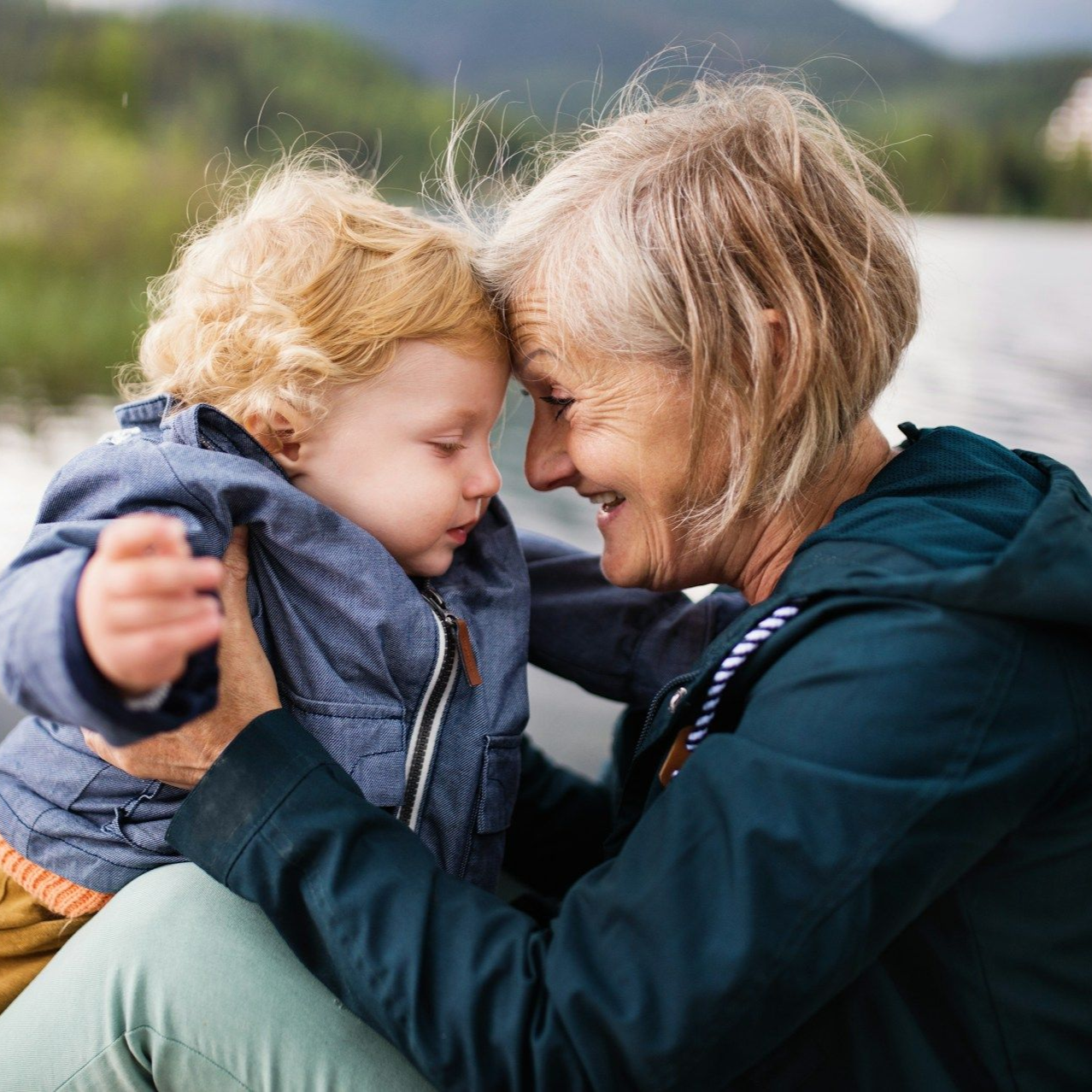 A woman and a young child touch foreheads, smiling. They sit outdoors near a body of water.