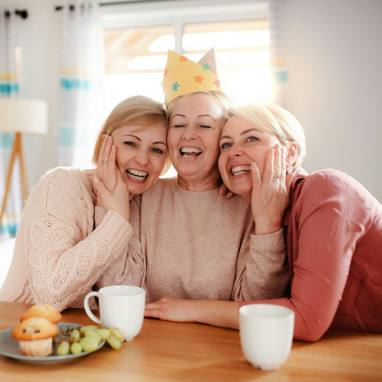 Three smiling women, one wearing a paper crown, at a table with mugs and a pastry; sunny indoor setting.