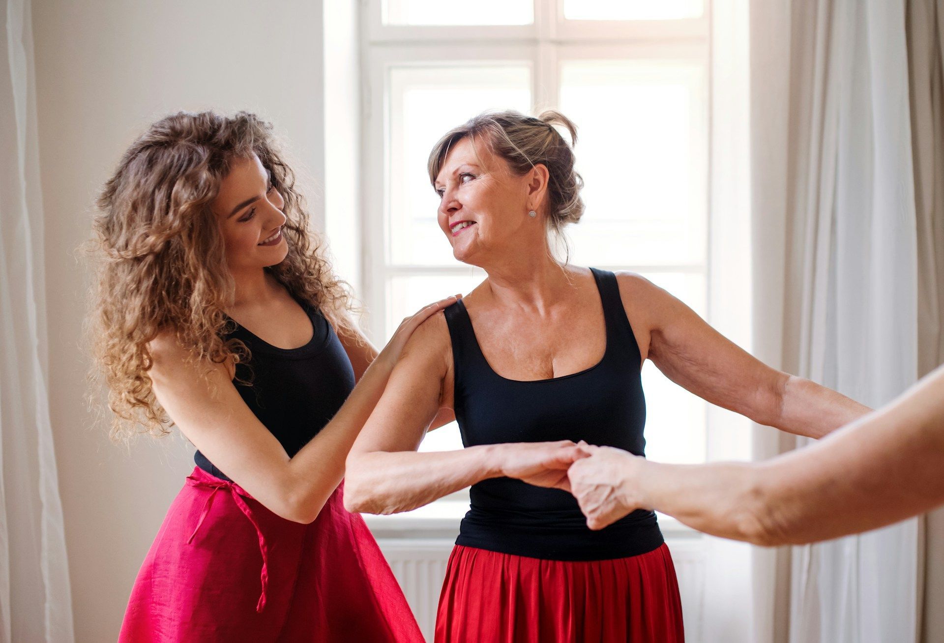 Two women in red skirts and black tops dance in a room with a window, smiling.