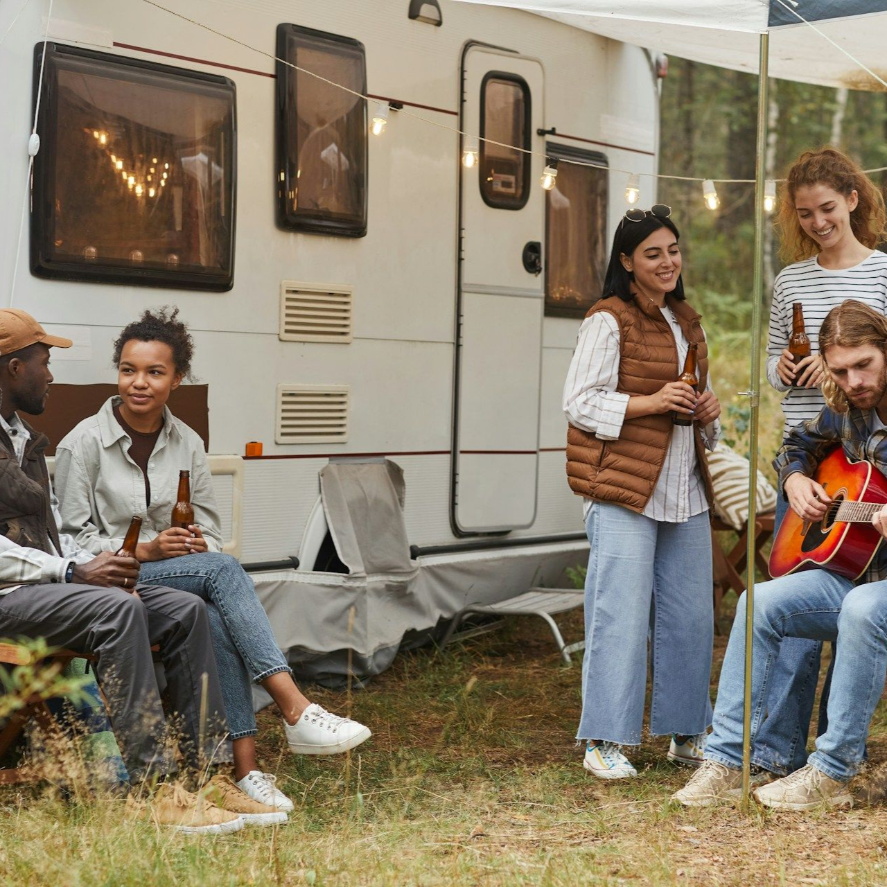 Group of friends camping, sitting, talking, and playing guitar by a trailer.