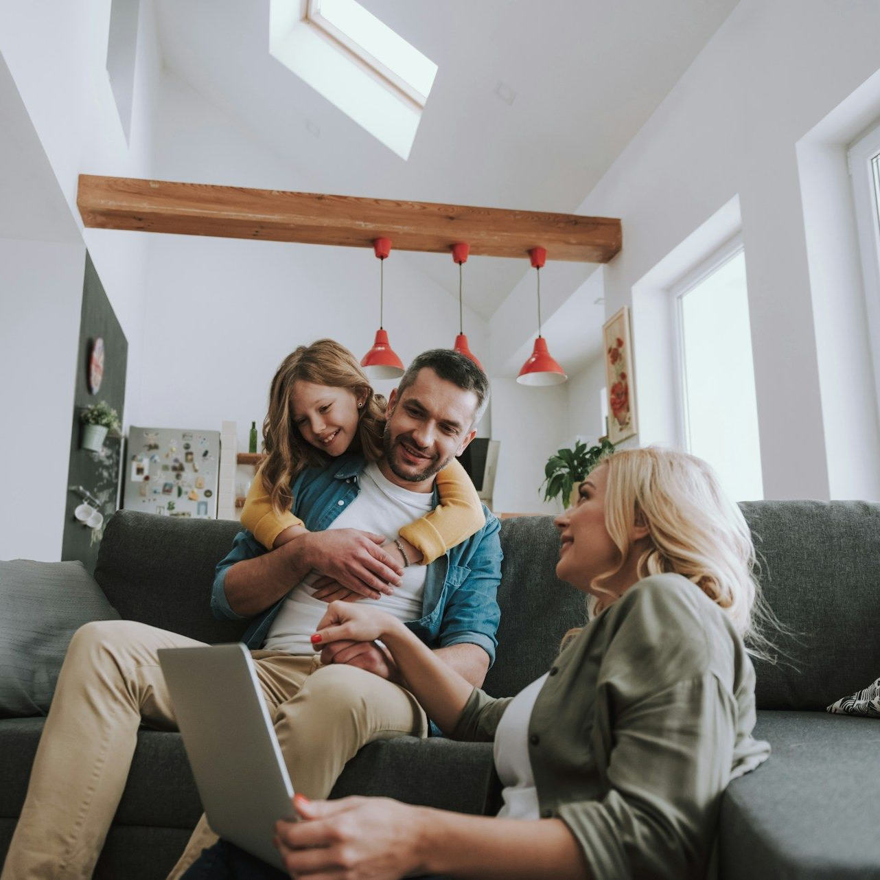Family in a modern living room: father embracing child, mother using laptop on couch.
