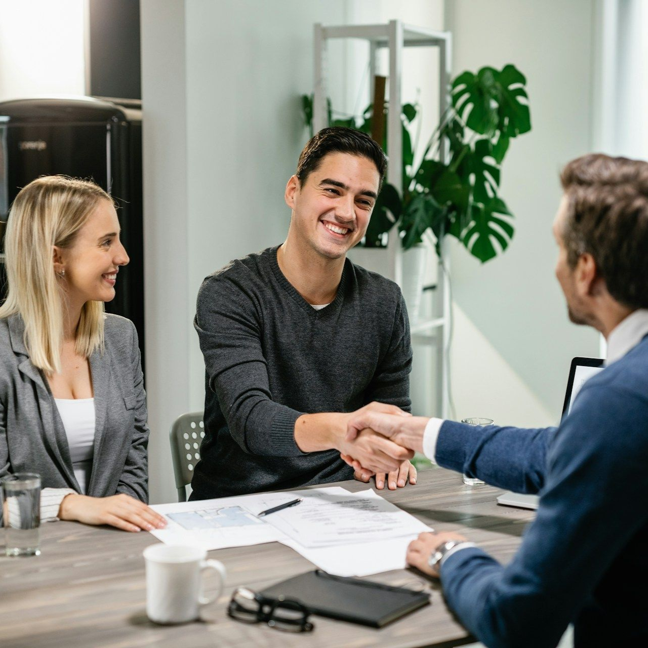 Couple shaking hands with a person at a table, smiling, in an office.