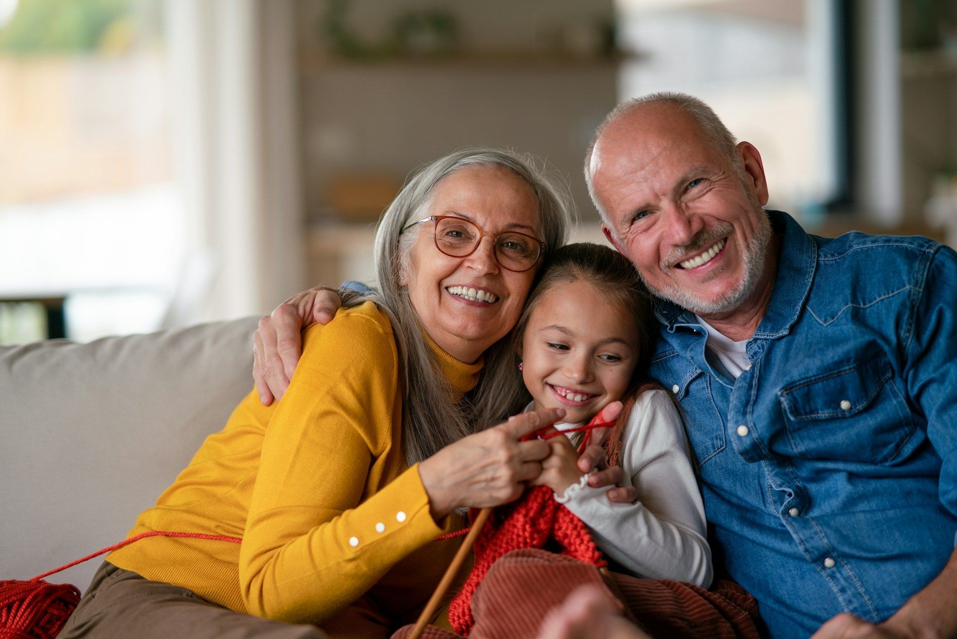 Grandparents with granddaughter knitting, smiling, on a couch.